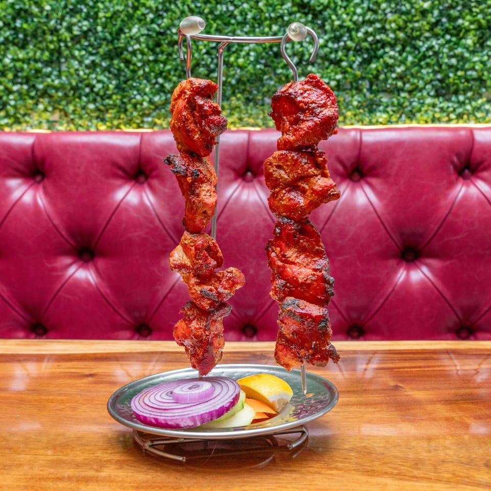 Skewers of grilled red-marinated meat hang over a plate of sliced onions, vegetables, and lemon on a restaurant table.