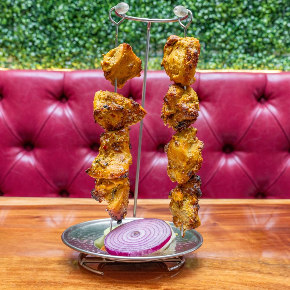 Spiced grilled meat skewers hang over a plate with sliced red onion, set on a wooden table against a burgundy banquette.