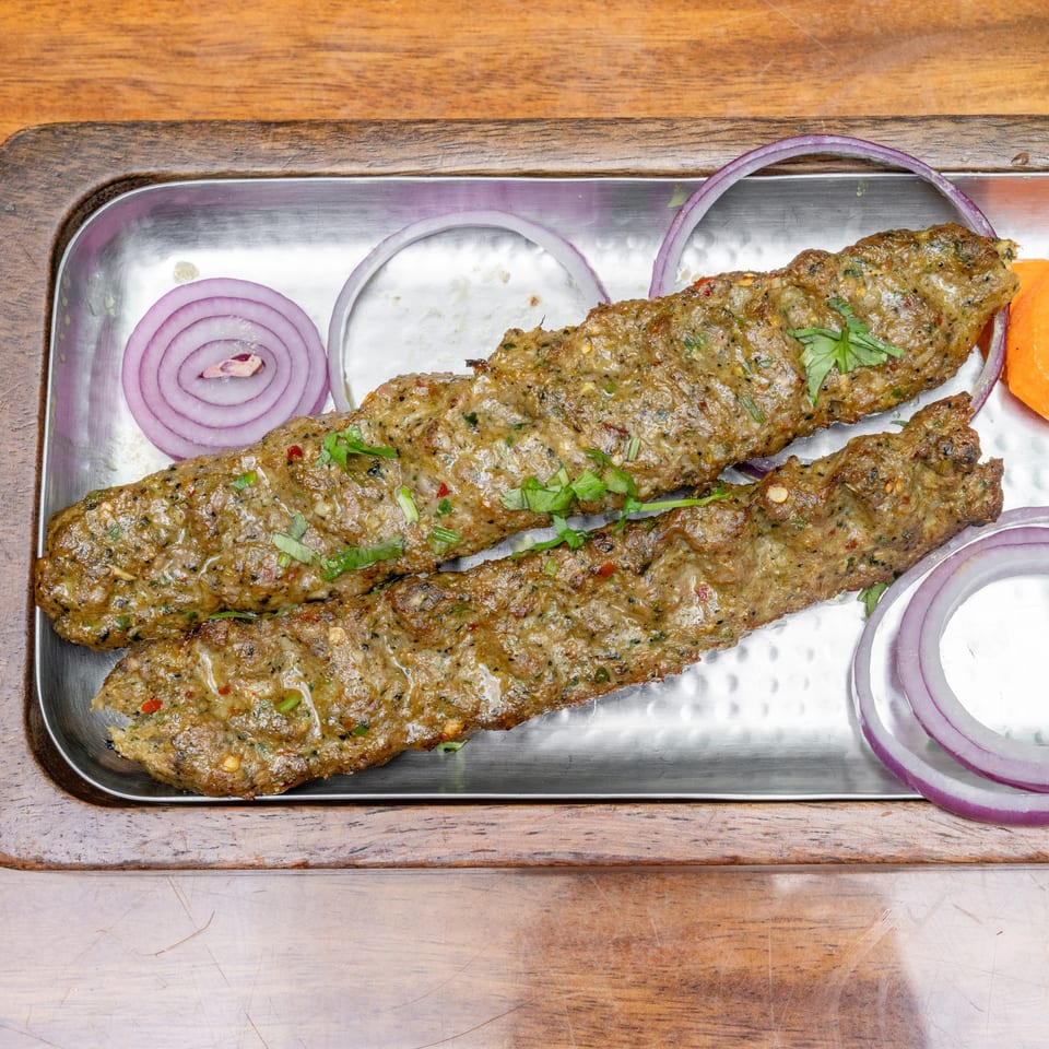 Three grilled seekh kebabs garnished with cilantro, red onion rings, carrot, cucumber, lemon, and yogurt sauce on a metal platter.