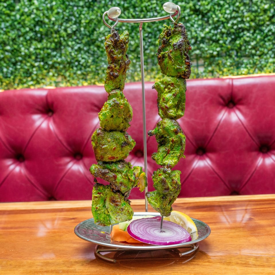Green-marinated grilled kebabs hang over a plate of red onion, lemon, and vegetables in a restaurant booth.