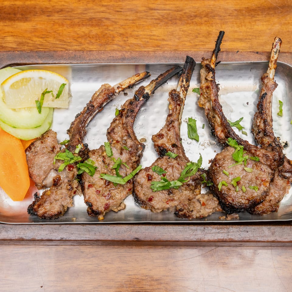 Grilled meat skewers and patties garnished with herbs, served with lemon, cucumber, and carrot slices on a metal platter.