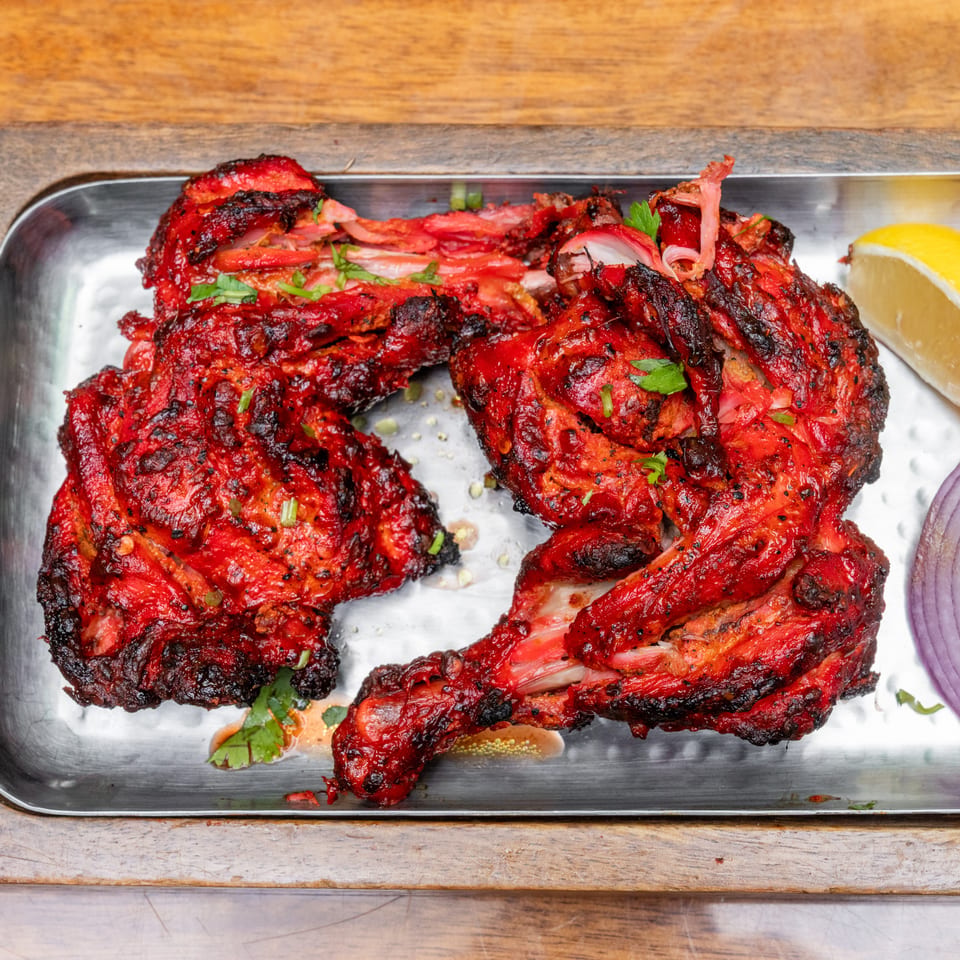 Charred red-spiced grilled chicken pieces garnished with herbs, served with lemon wedges, tomato, and sliced red onion on a metal platter.